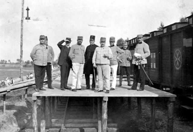 Najprawdopodobniej ten pociąg szpitalny odwiedził w listopadzie 1914 r. Limanową. Na burtach wagonów doskonale widoczne znaki Czerwonego Krzyża i charakterystyczne, ośmiokończaste Krzyże Maltańskie. Na drewnianej rampie grupa oficerów i cywili z personelu szpitala pozuje do zdjęcia. Na drugim planie, z lewej, drewniana pochylnia służąca do transportu rannych. W porannym słońcu wygrzewa się tam druga grupa obsady pociągu. Na uwagę zasługuje ciekawa lampa wisząca na wysokiej żerdzi. Zapewne postój pociągu zaplanowano w strefie przyfrontowej, w oczekiwaniu na rannych. Na awersie tej unikalnej fotografii widnieje tylko napis Galizien. Brak danych umożli- wiających dokładną lokalizację, ale równinne tereny wskazują na Galicję Wschodnią.